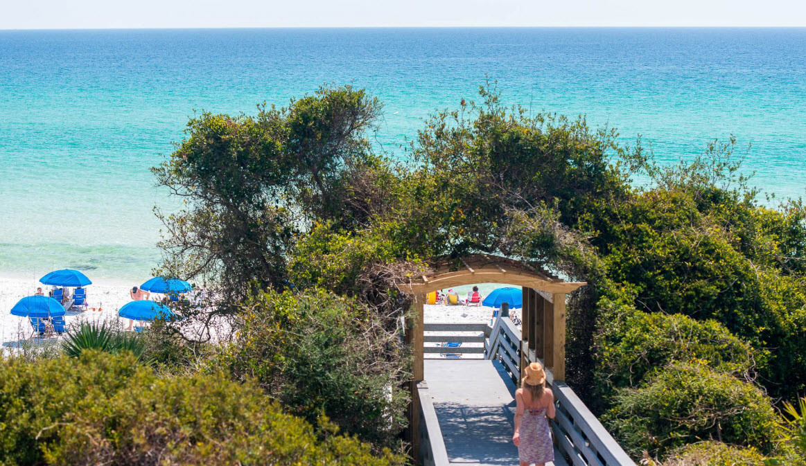 Woman walking a beach boardwalk beneath an arch of greenery