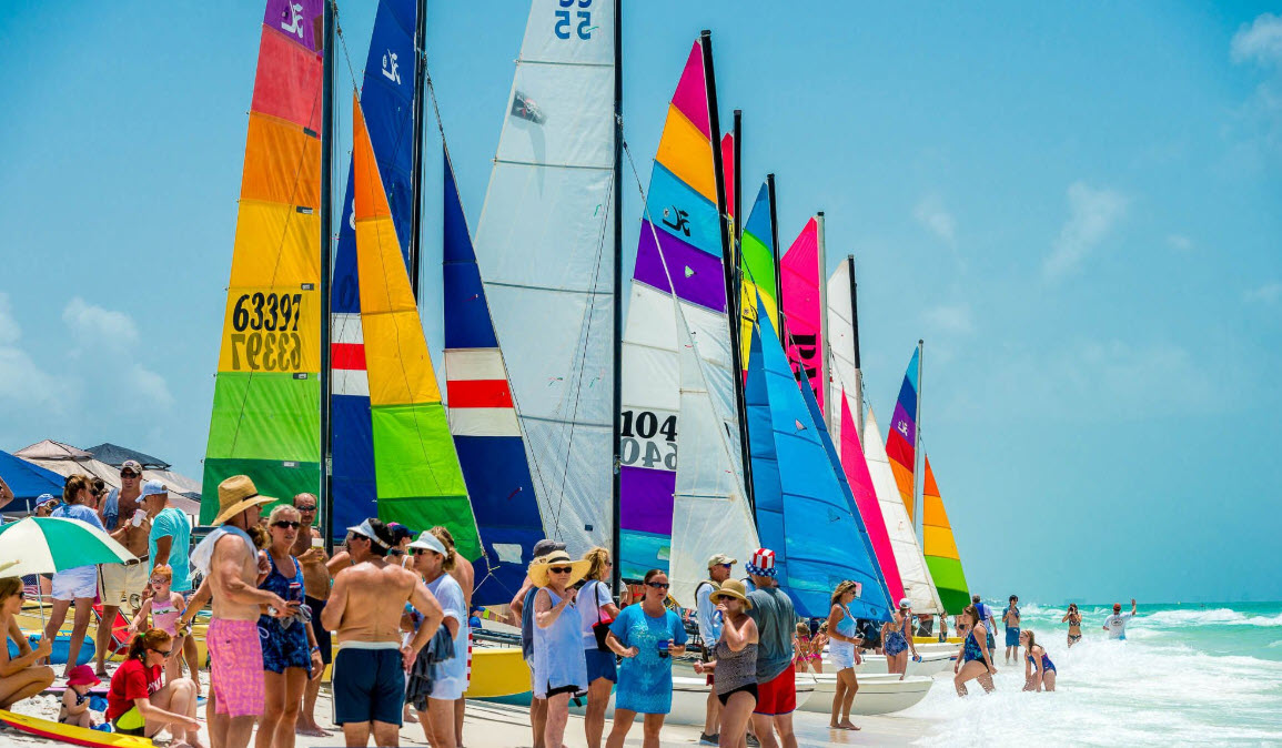 Families gathered among colorful sailboats on the beach