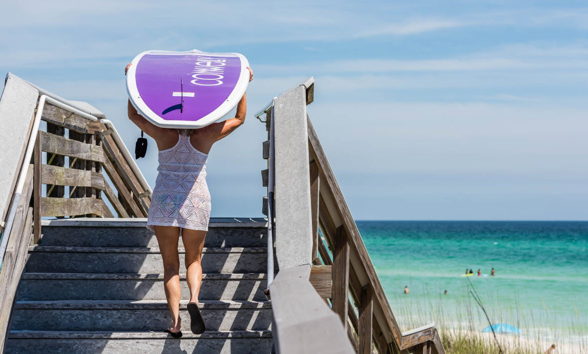 Person carrying a purple paddleboard up beach stairs